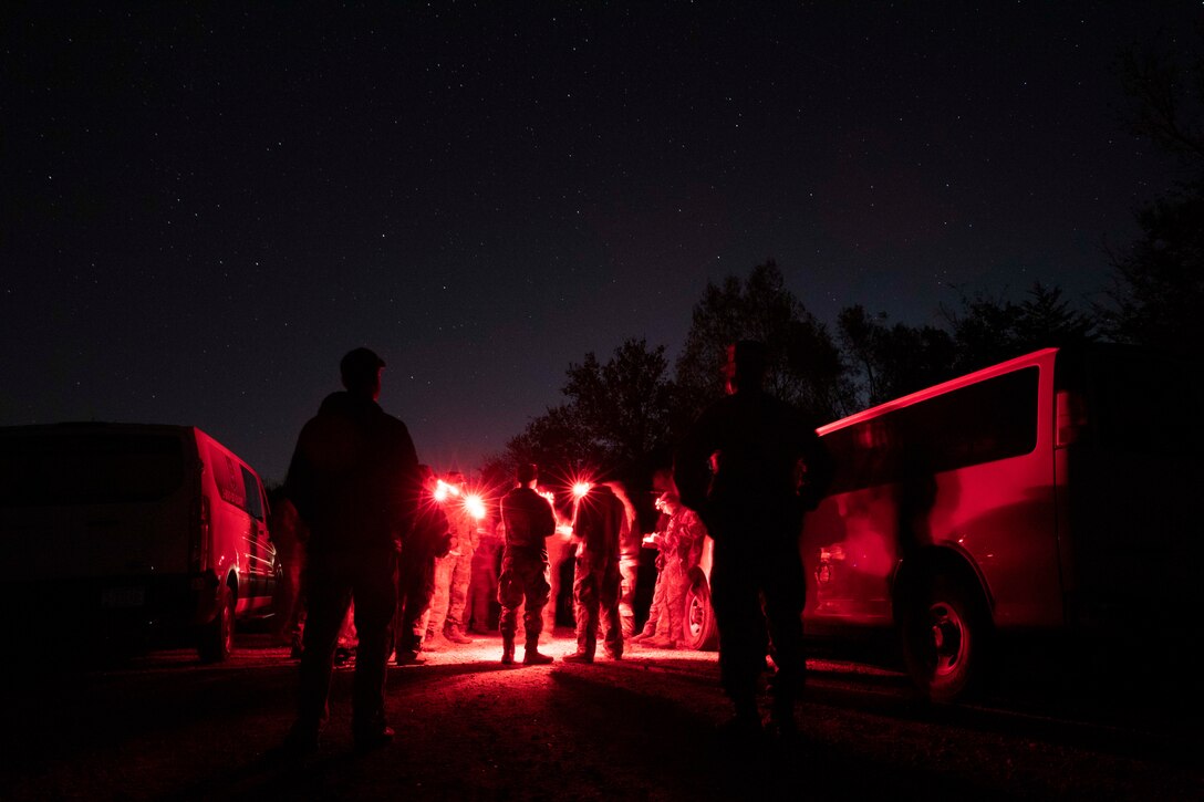 Soldiers gather in a circle near vehicles under a starry night sky illuminated by red lights.