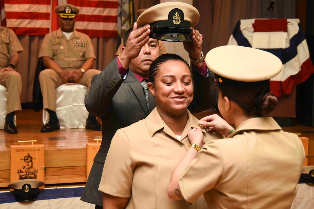 A man places a hat on top of a sailor’s head as another sailor pins a pin on the sailor’s uniform.