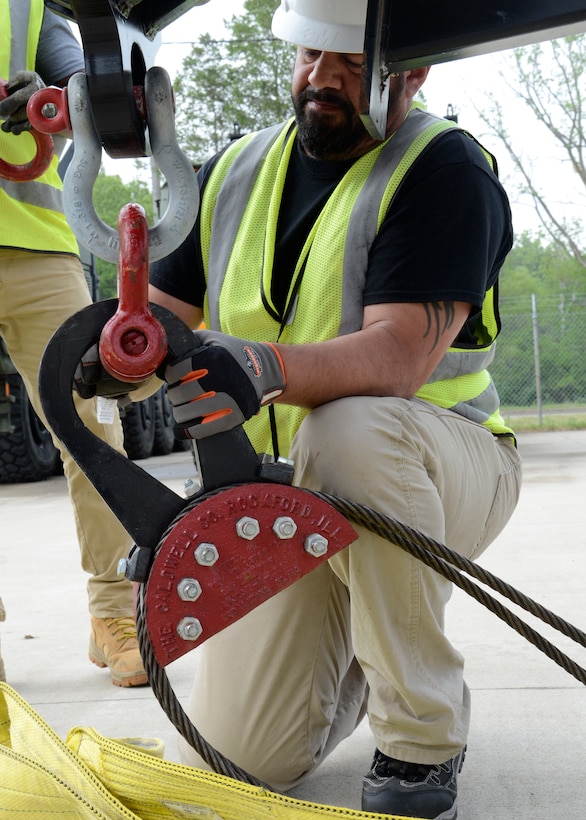 A man in safety vest and hard hat works with a heavy cable and hook as part of a rigging system.