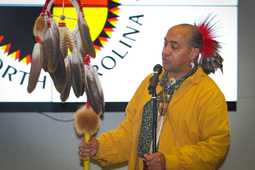 A member of the Lumbee Nation displays artifacts in front of a large screen.