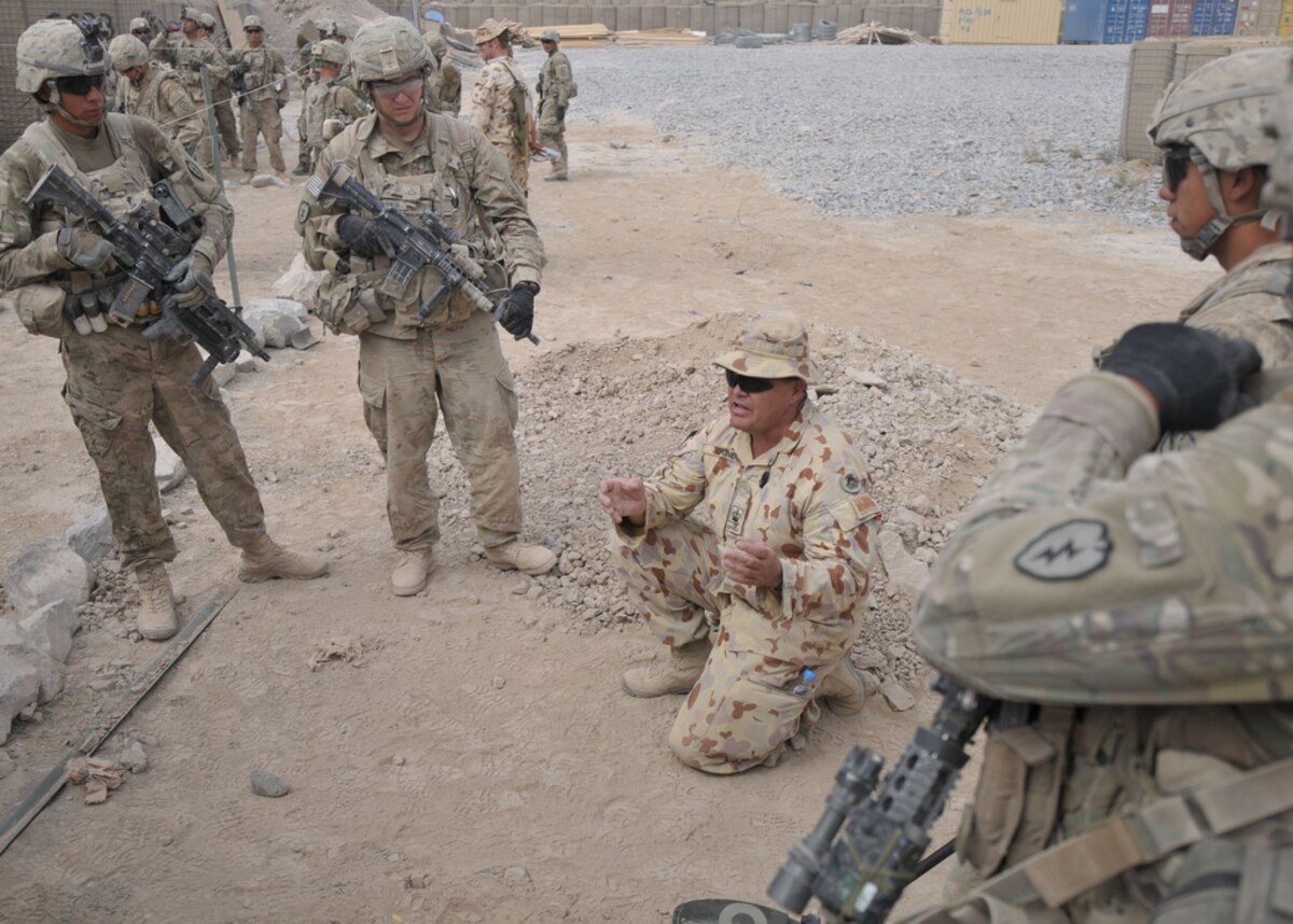 A man in a military uniform kneels on the ground. Others stand around him.