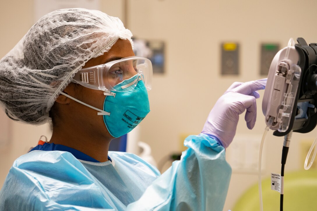 A military medical technician wearing a mask and gloves uses a medical device.