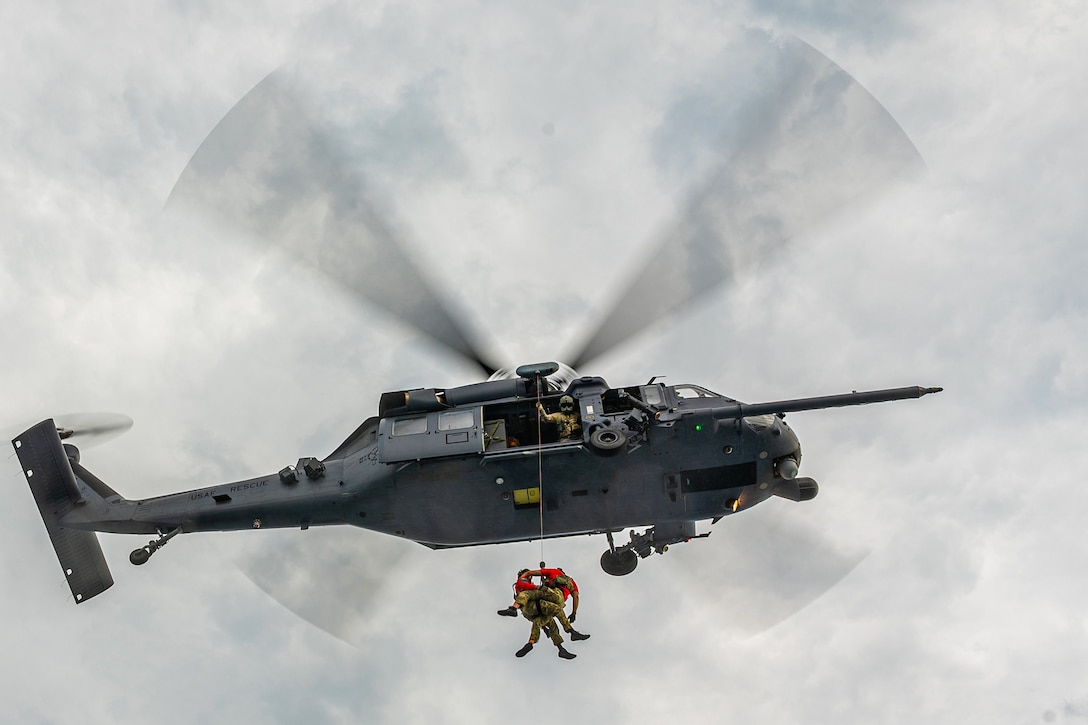 Two airmen hover by a rope beneath an airborne helicopter.