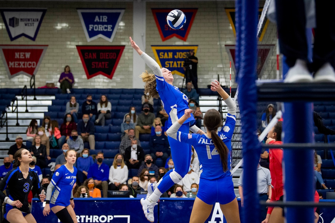 An airman, surrounded by fellow athletes and spectators in the stands, reaches for a volleyball off the ground.