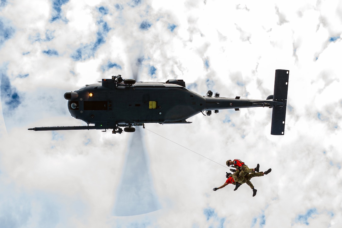 Two airmen hover from an airborne helicopter by rope.