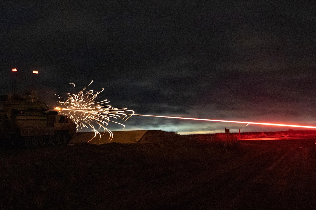Gold and red sparks fly as a round fires from a tank in an open field at night.
