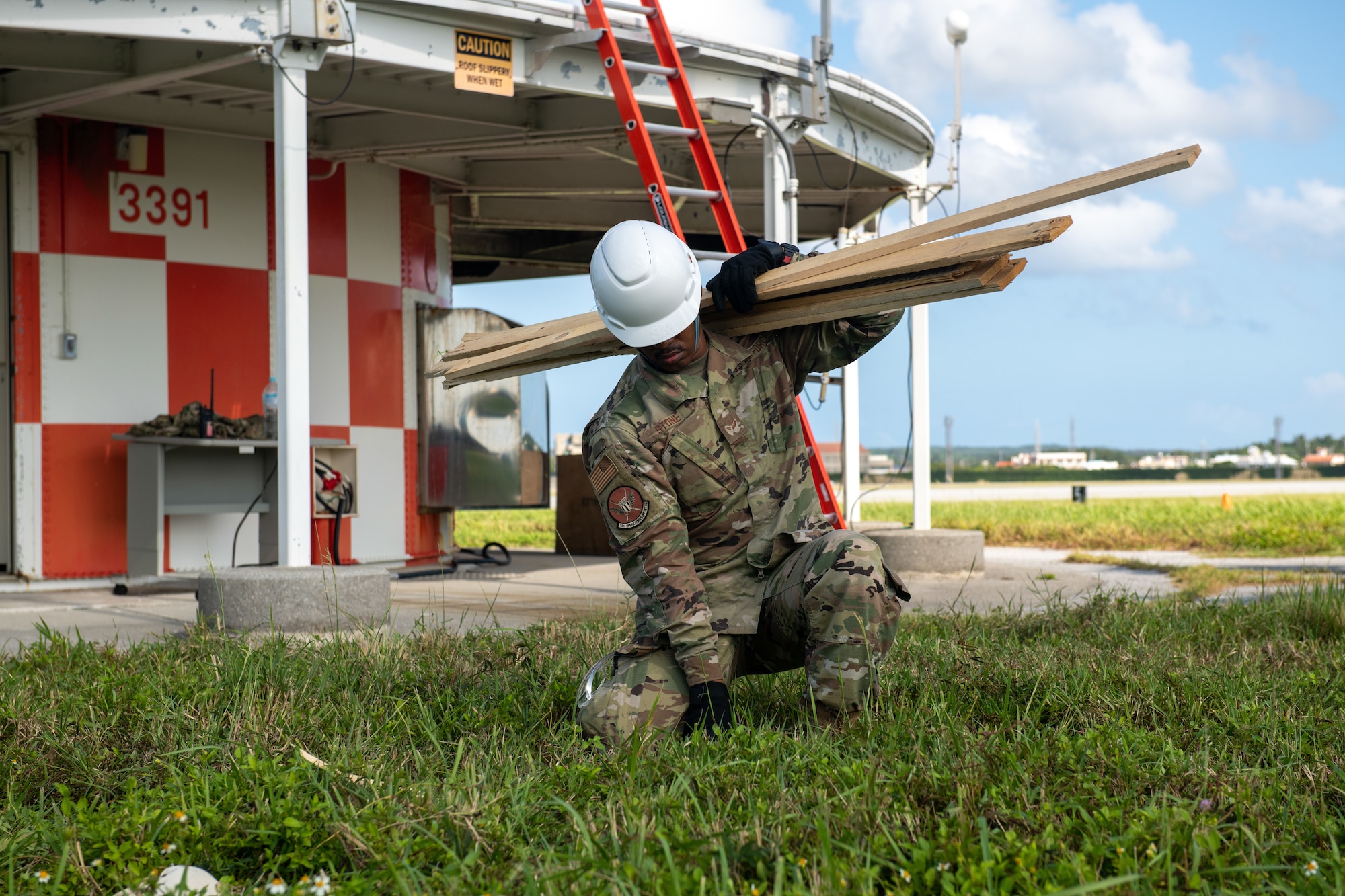 An Airman picks up wooden planks in front of an orange and white building