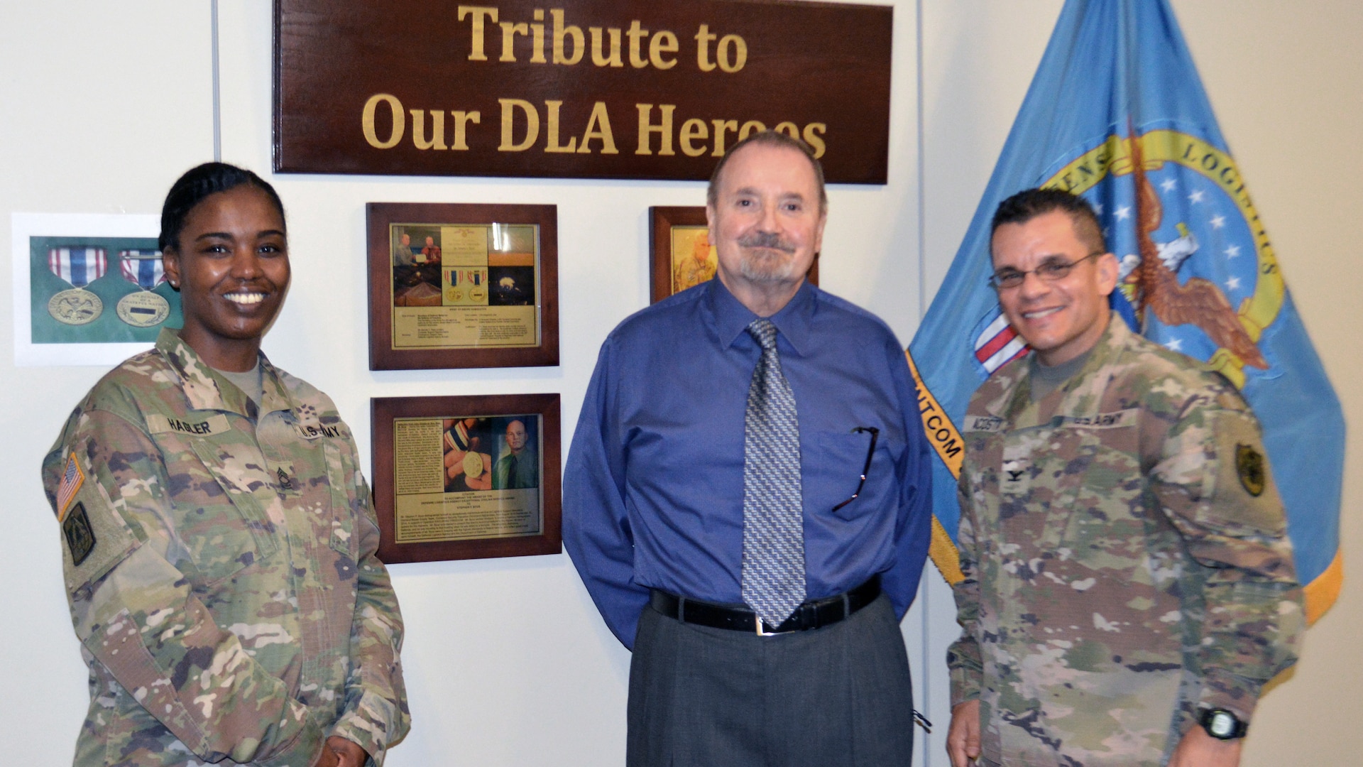 One woman and two men stand in front of a white wall with plaques behind them.