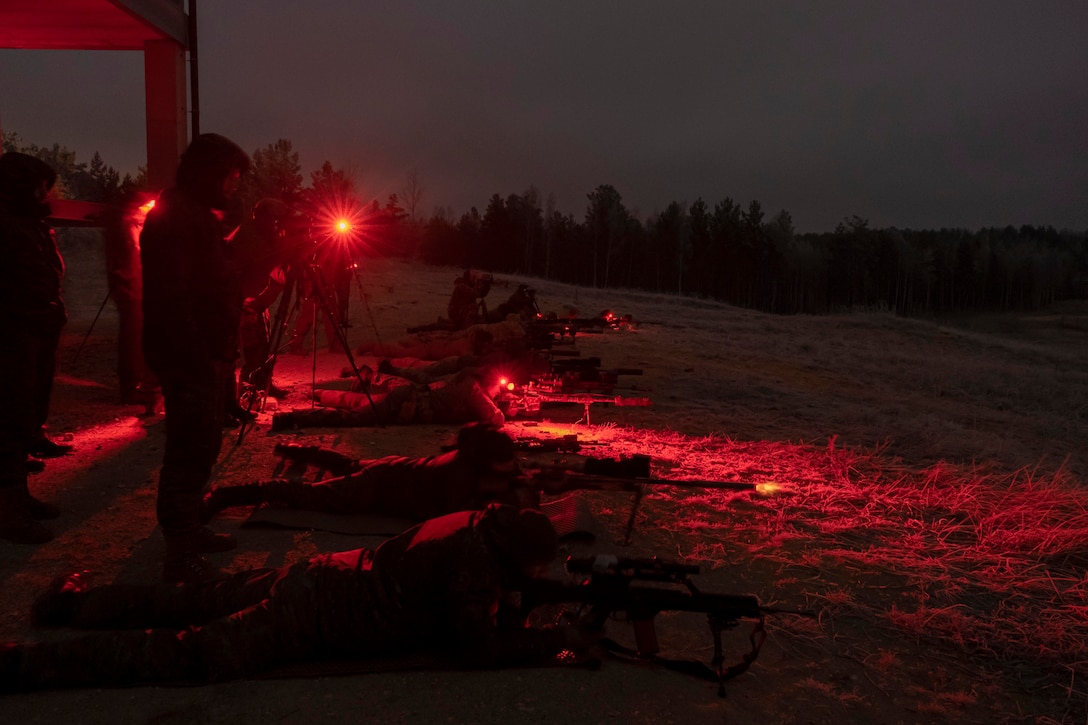 Soldiers laying on the ground fire at targets illuminated by red light.
