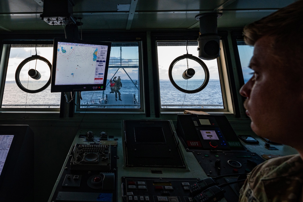 A soldier watches out of a ship's window as soldiers conduct hoist operations.