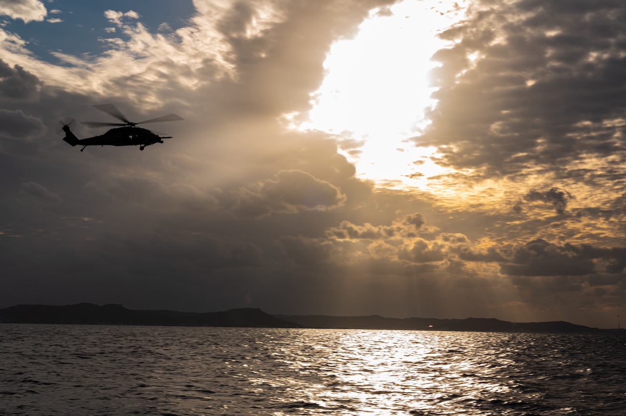 A helicopter flies over a body of water under a sunlit sky.