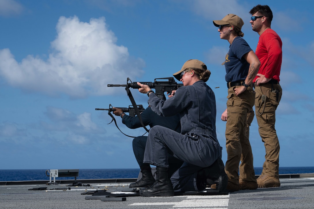 Sailors kneel on the flight deck of a ship and fire rifles.