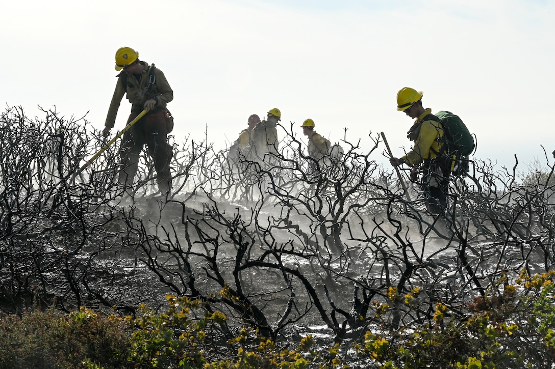 A group of airmen wearing firefighting gear search through a smoky burned area.