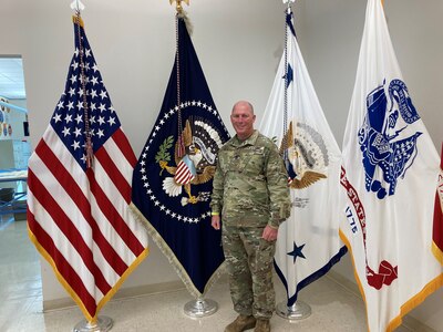 Male Army officer poses in front of flags.