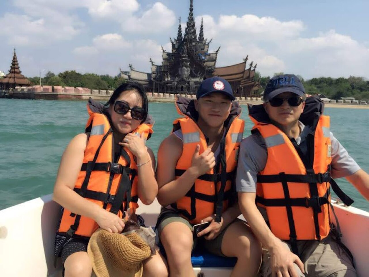 Three people wearing life preservers pose for a photo in a boat, with an ornate Thai building in the background.