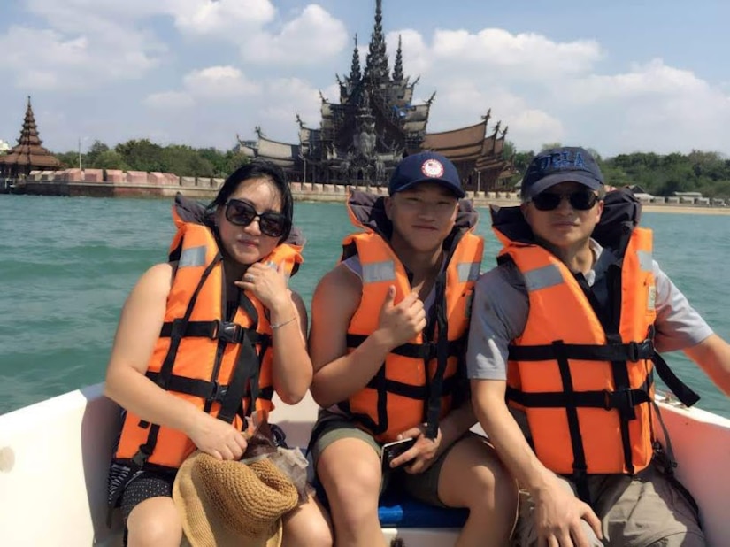 Three people wearing life preservers pose for a photo in a boat, with an ornate Thai building in the background.