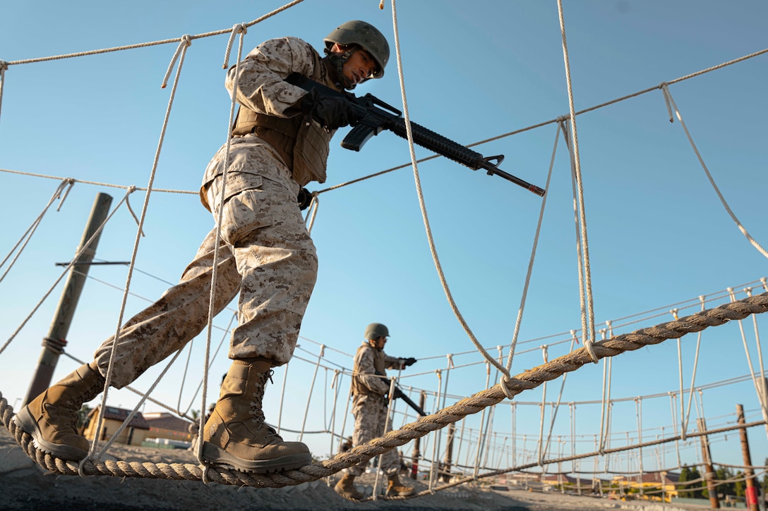A Marine Corps recruit walks across a rope while carrying a weapon.