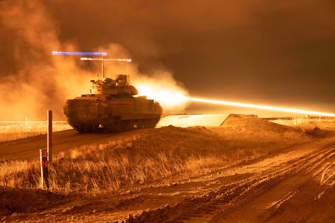 An armored vehicle fires in a field illuminated by orange light.