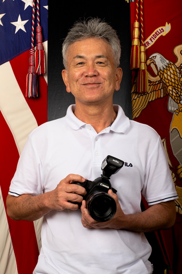 Naoshi Higa, a photo laboratory technician with the Marine Corps Installations Pacific Combat Visual Information Center (CVIC), poses for a photo with his Canon 5D Mark III still camera at the CVIC, Camp Foster, Okinawa, Japan Sept. 15, 2021. Mr. Higa has worked for the CVIC, formerly Marine Corps Bases - Japan combat camera, for 36 years assisting service members stationed on Okinawa in updating and capturing official photos to be used for passports, command boards, promotion boards, tattoo documentation, and other military requirements. (U.S. Marine Corps photo by Cpl. Ryan H. Pulliam)