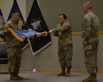 Three military members in uniform stand on a stage. The female member in the center holds a flag straight in front of her as she spins it along the pole, assisted by the male military member in the left of the photo as the male to the far right oversees the flag retirement.