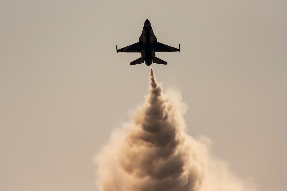 A fighter jet flies in a grey sky with smoke trailing behind.