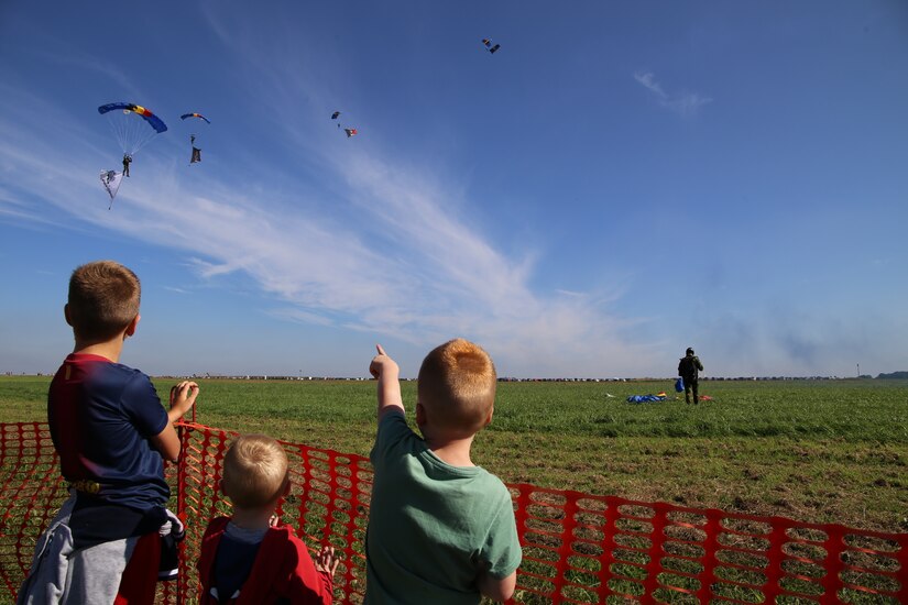 Three children look up at people descending in the sky wearing parachutes.