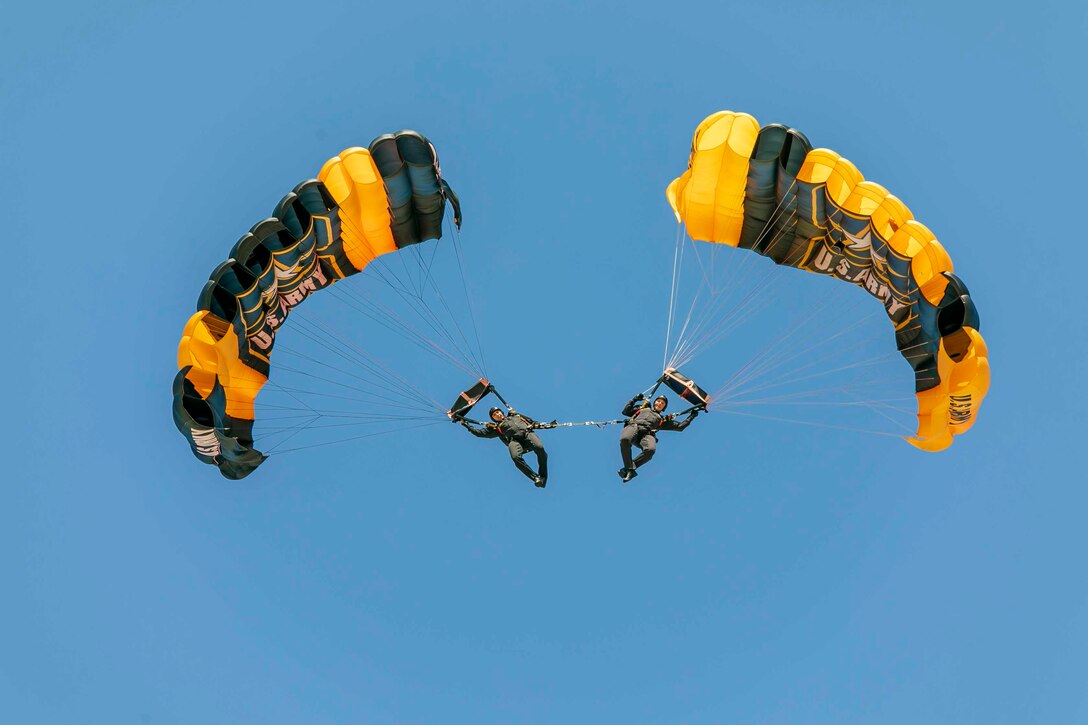 Two soldiers connected by a strap descend in the sky using parachutes.
