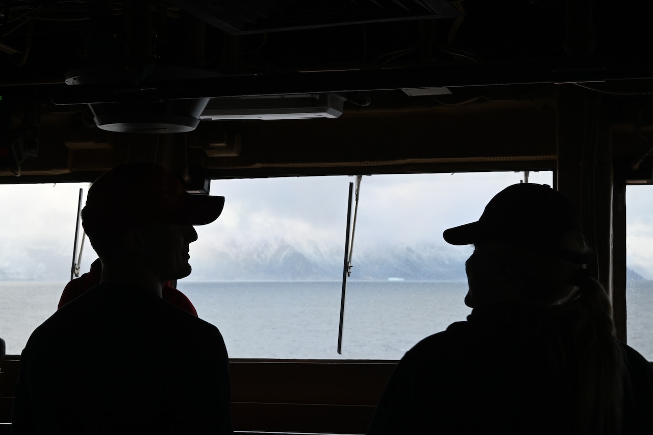 Two people are silhouetted against a window looking out on the ocean and clouds.