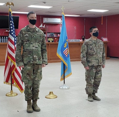 Two military men stand at attention during a change of command ceremony with flags in background.