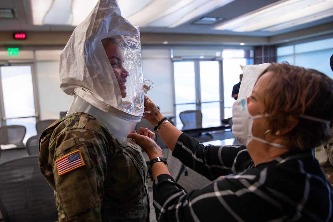 A female soldier stands as a woman wearing a face mask assists her in a respirator fit test.
