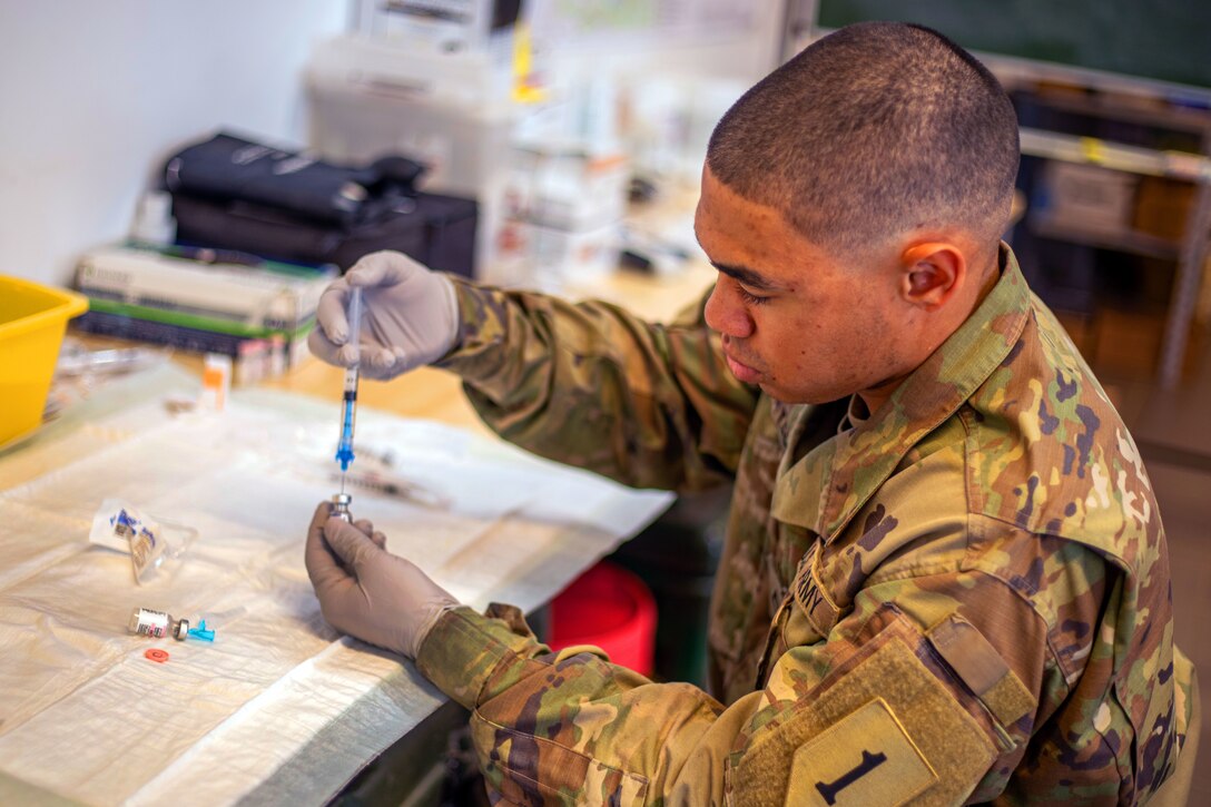 A soldier wearing gloves holds a syringe and small bottle.
