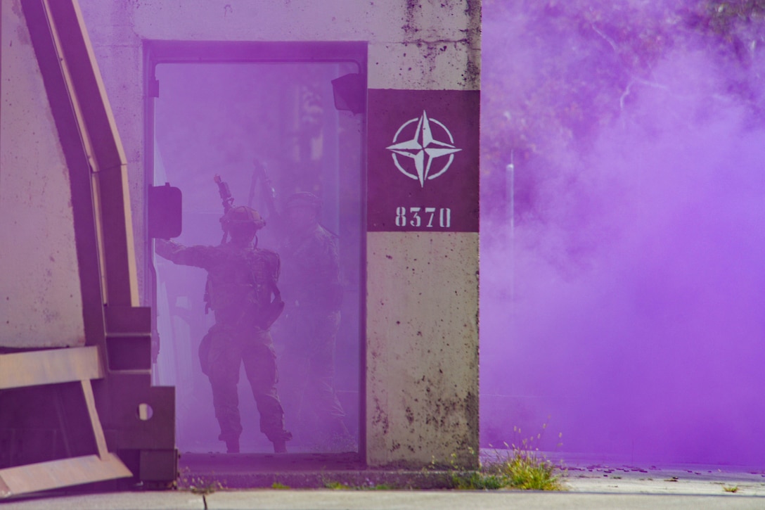 Two airmen holding weapons talk in a warehouse-like area surrounded by clouds of purple smoke.