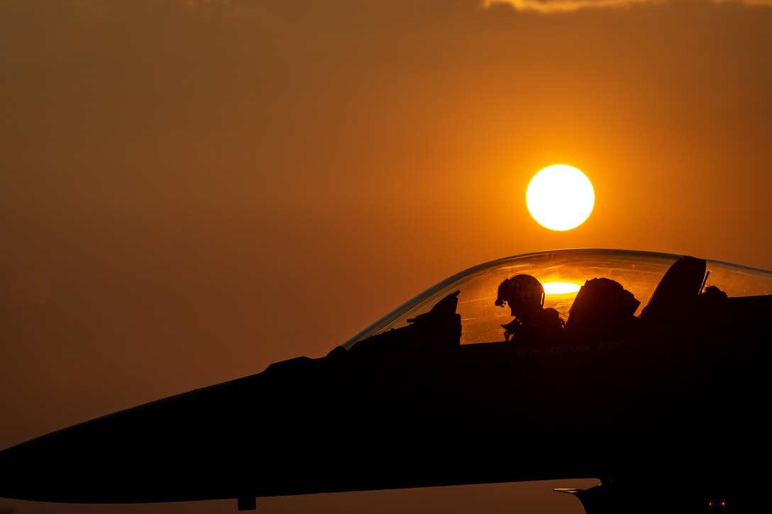 An airman sits in the cockpit of an aircraft under a sunlit sky.