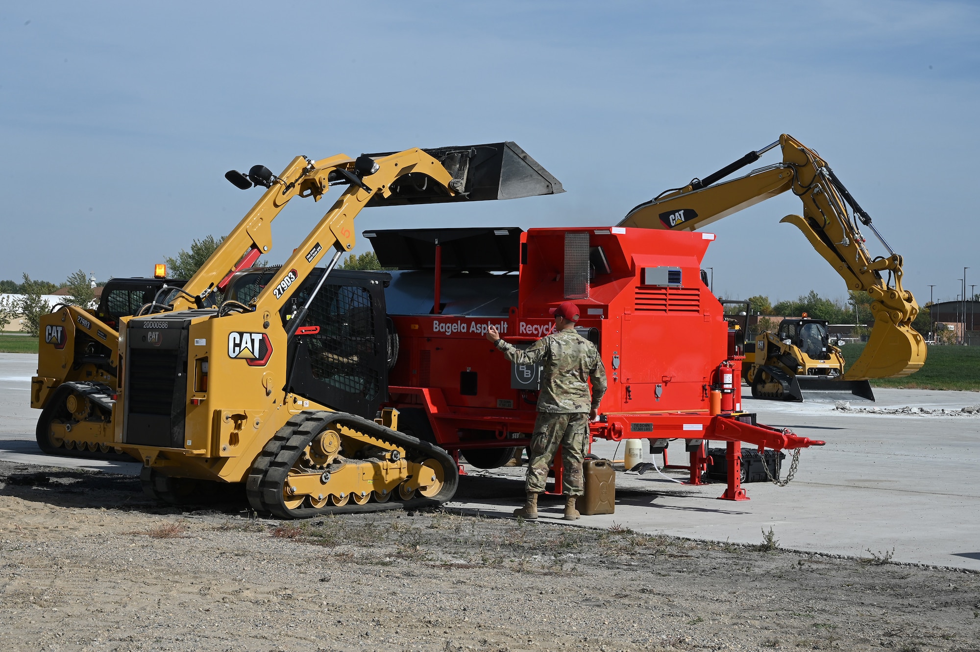 A military member directs a skid steer operator as he loads asphalt material into a large machine called an asphalt recycler to be reformed into patching material used to patch holes on a large concrete training runway at the North Dakota Air National Guard Regional Training Site, Fargo, N.D., Sept. 29, 2021.