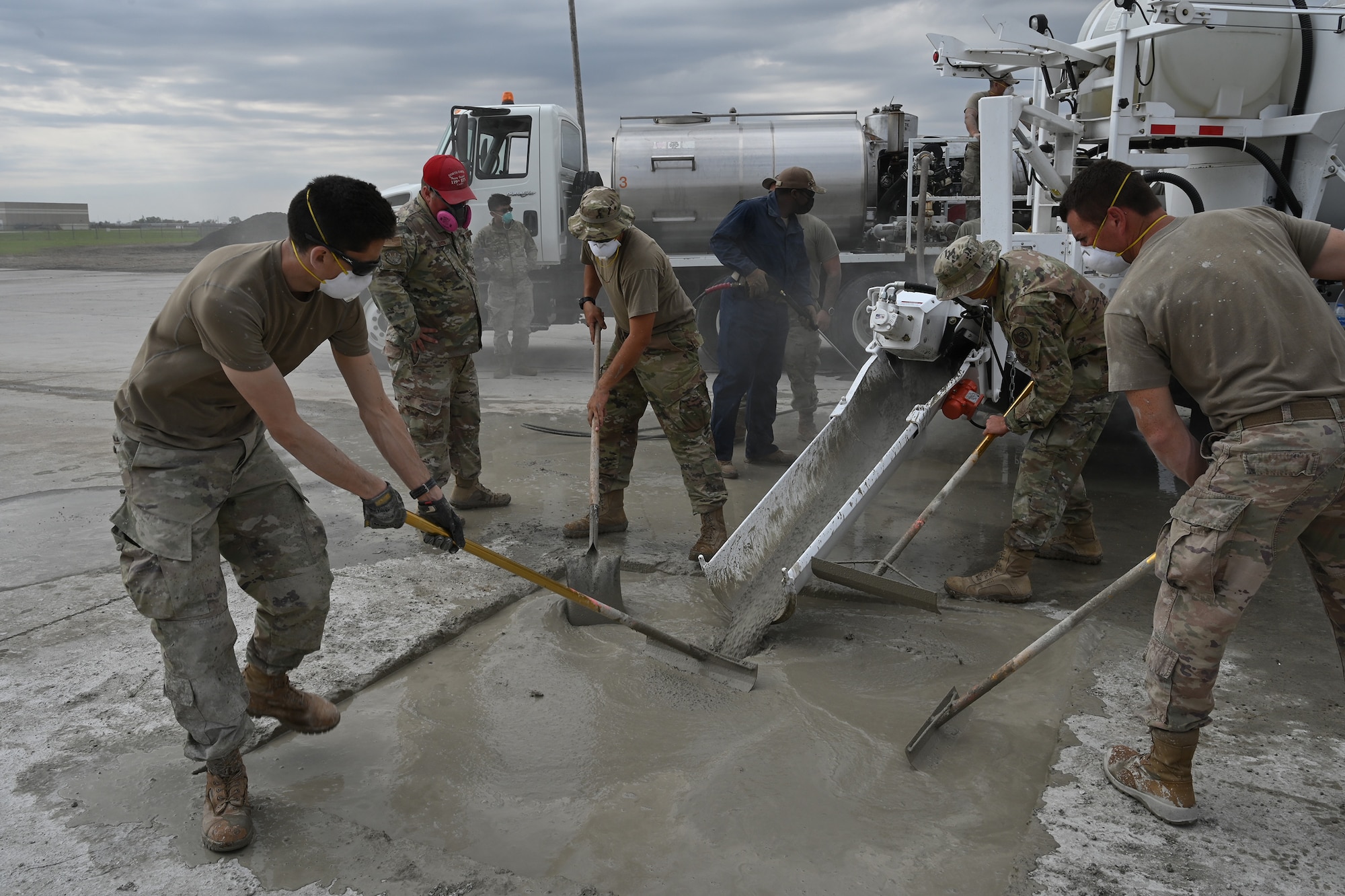 Four military members smooth quick-set concrete with rakes in a square hole in an effort to patch the hole for training at the North Dakota Air National Guard Regional Training Site, Fargo, N.D., Sept. 30, 2021. The concrete patching material is coming down a metal shoot from a machine called a volumetric mixer.