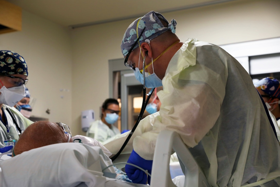 A soldier wearing a face mask, gloves, scrub cap and medical gown cares for a COVID-19 patient while another medical tech wearing a face mask, scrub cap and medical gown assists while several medical techs observe.