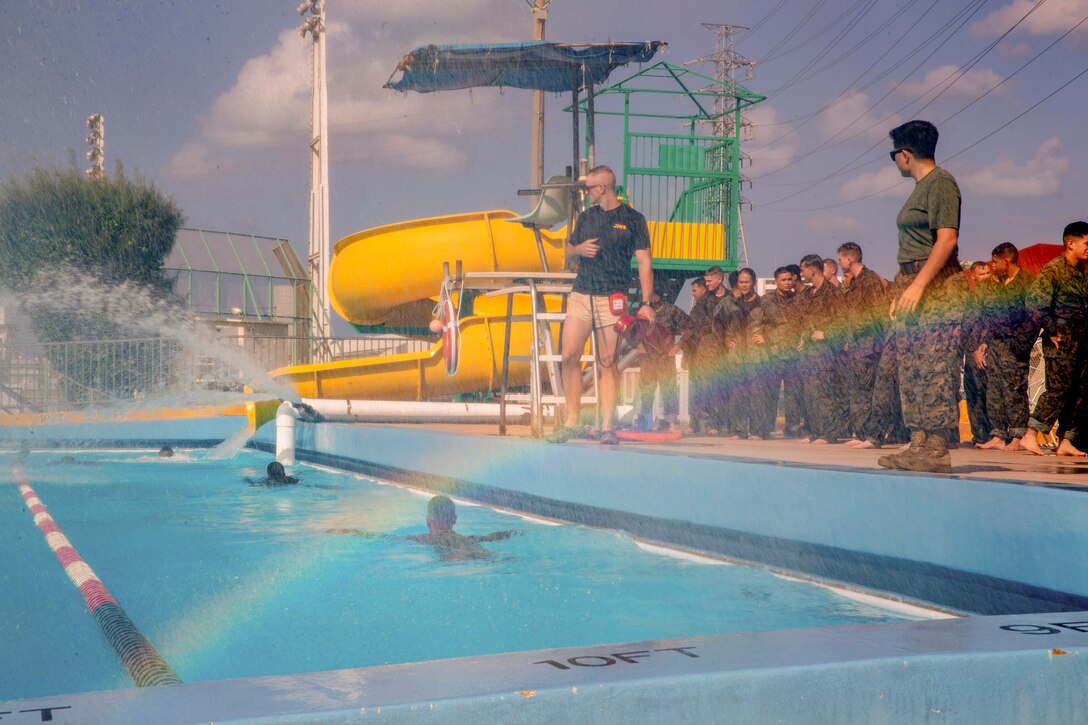 Marines swim laps in a pool covered by a rainbow as fellow Marines watch.