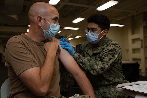 Hospitalman Christopher Ramirez administers a COVID-19 vaccine to Lt. Cmdr. George Stegeman, John F. Kennedy’s psychological officer, during a joint vaccination evolution