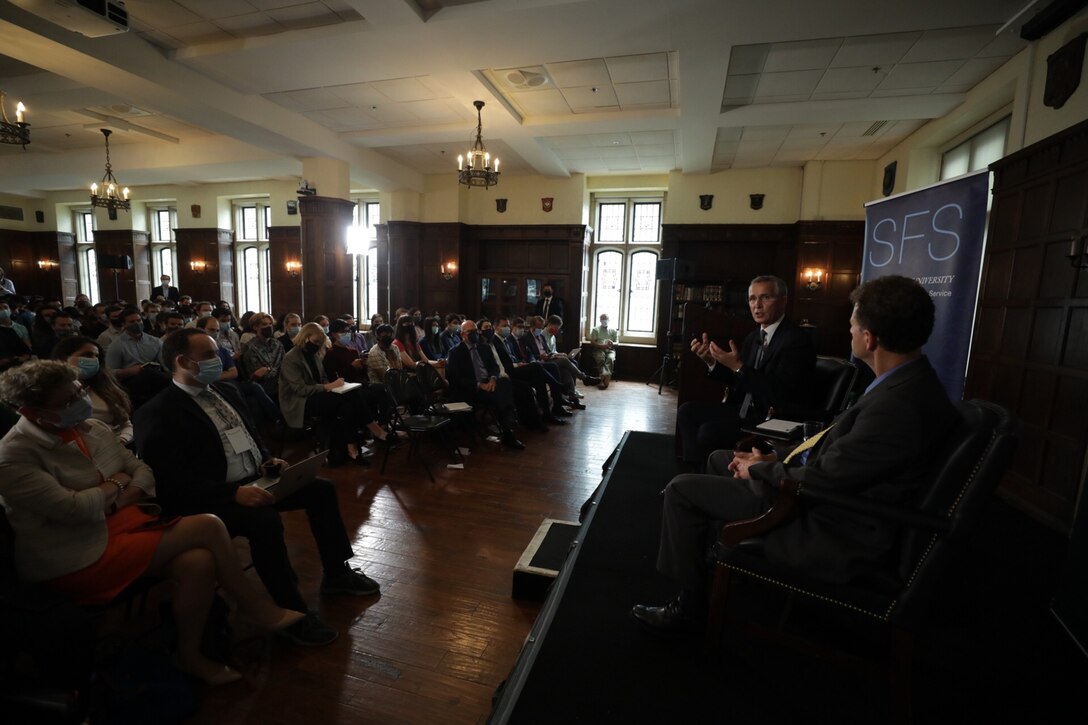 Two men sit on a stage in front of a roomful of people.