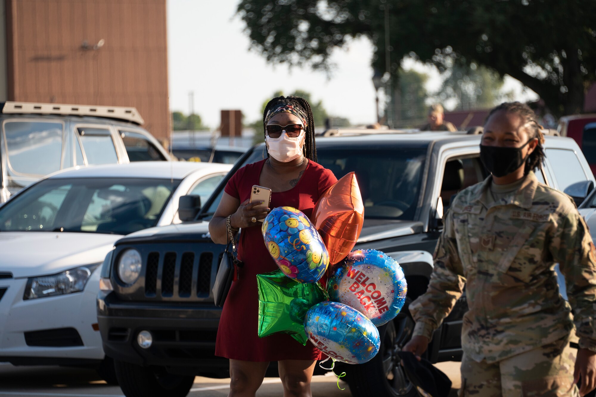 A woman stands holding five balloons, a woman in uniform walks forward.