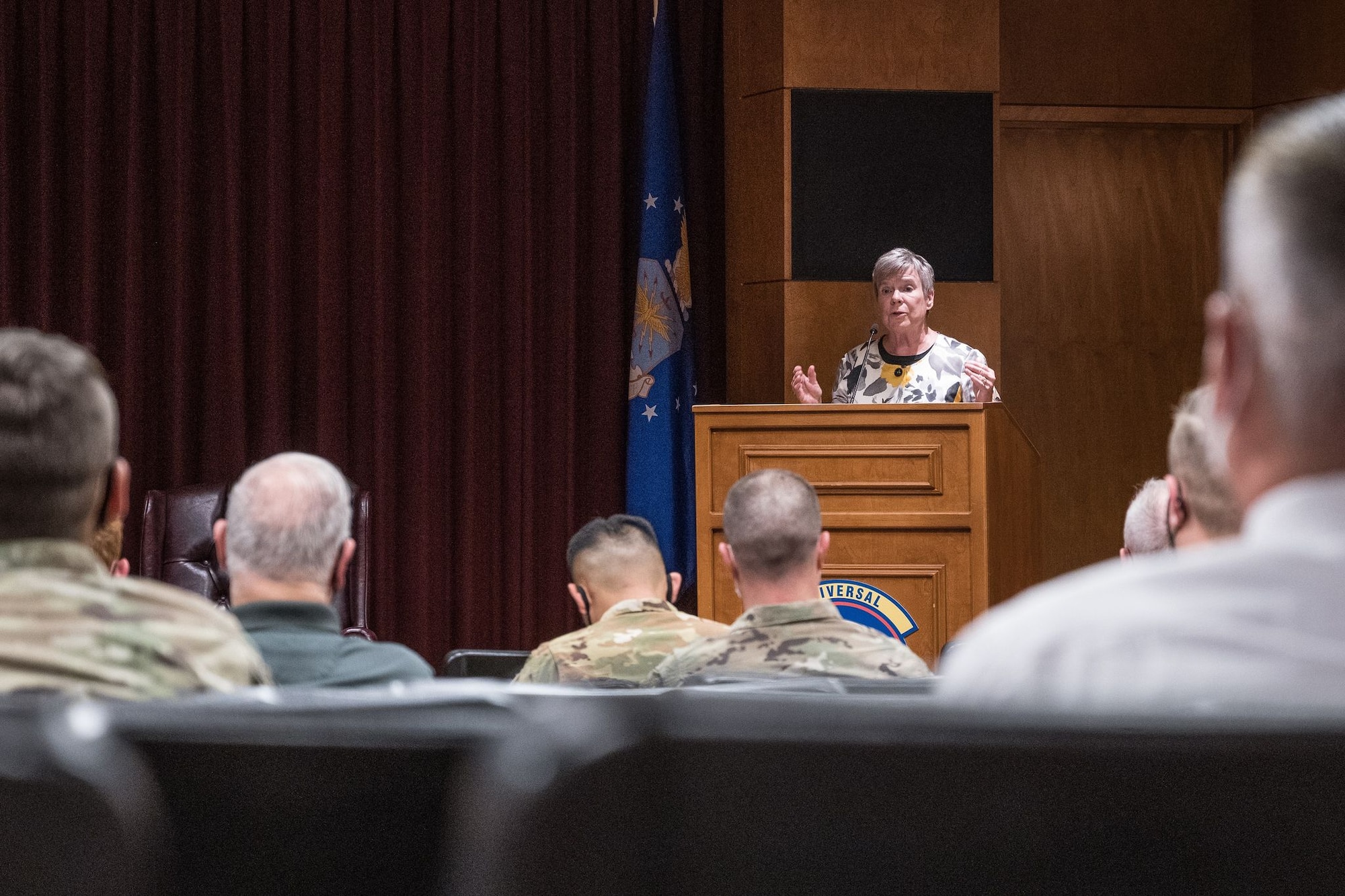 A woman stands at a podium in front of a crowd of uniformed service members in a conference room