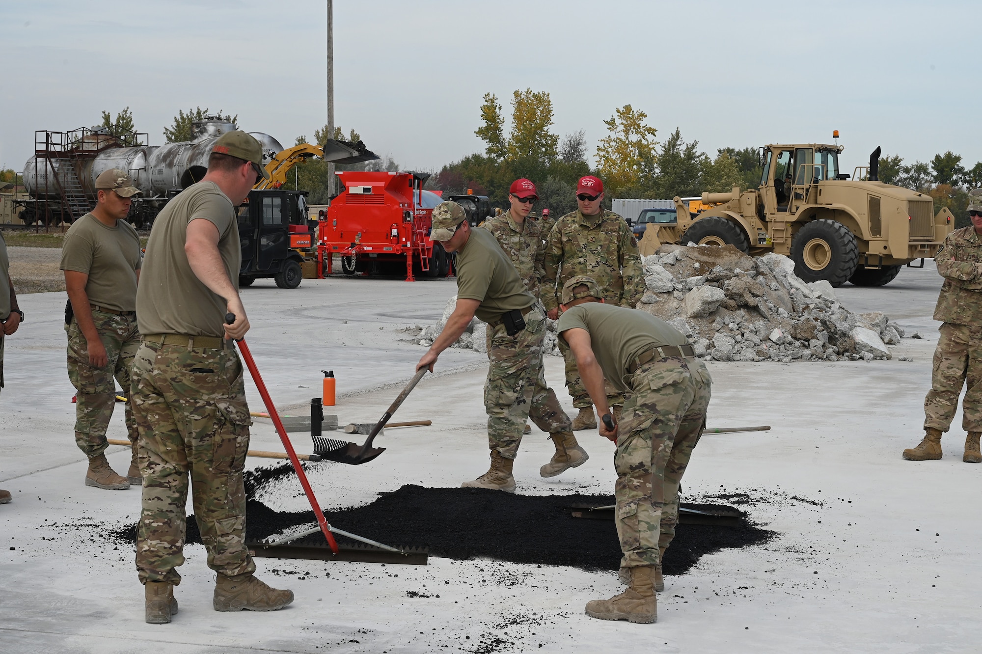 Three military members use rakes to smooth out asphalt material used to patch a square hole on a concreate training runway as several other personnel watch at the North Dakota Air National Guard Regional Training Site, Fargo, N.D., Sept. 29, 2021.