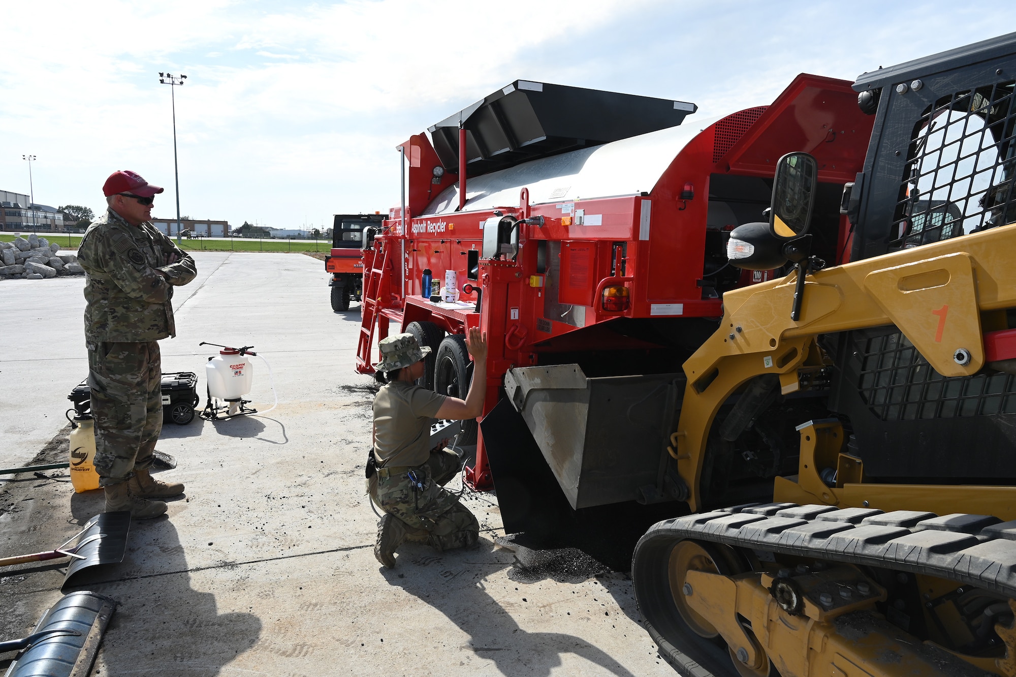 Two military members operate a large red machine as called an asphalt recycler as it heats asphalt material for use patching holes on a large concrete training runway at the North Dakota Air National Guard Regional Training Site, Fargo, N.D., Sept. 29, 2021.