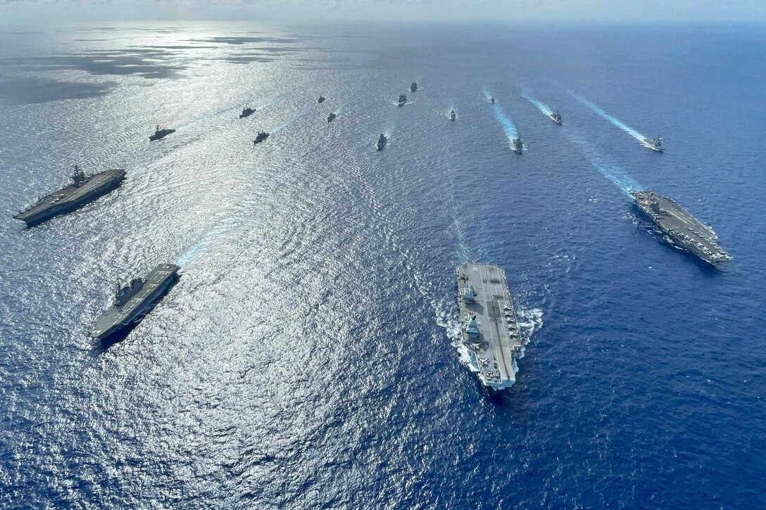 A large group of military ships move through the ocean in formation.
