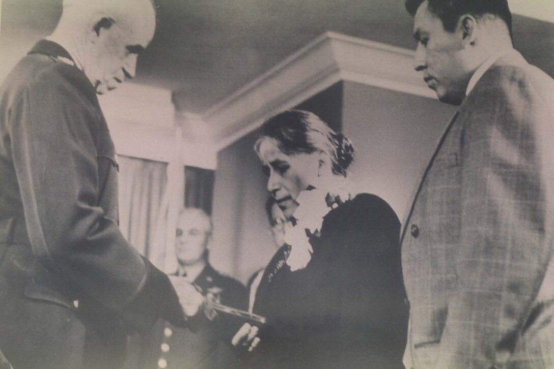A woman looks down at something handed to her by a tall man. Another man stands beside her.