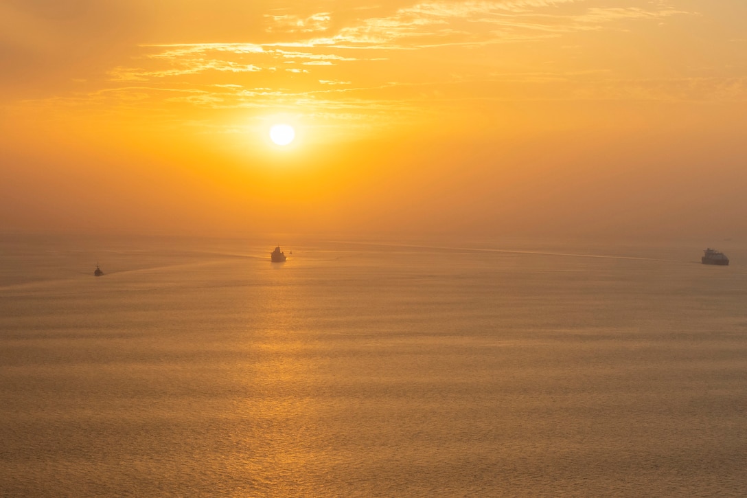 Three ships transit a body of water under a sunlit sky.