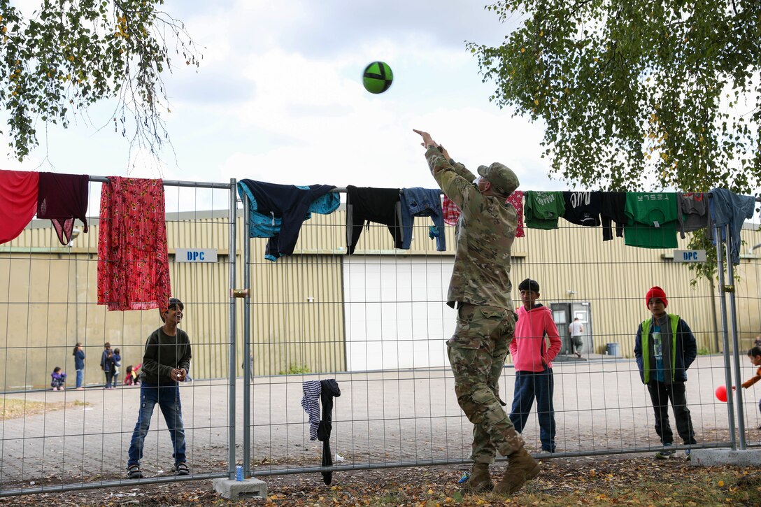 A soldier throws a ball over a metal fence to a child.