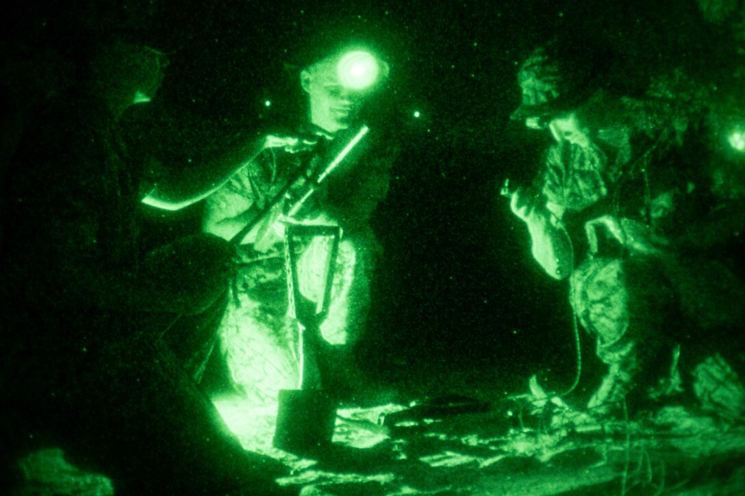 Three Marine Corps recruits hold weapons and gather in a circle as seen through night vision goggles.
