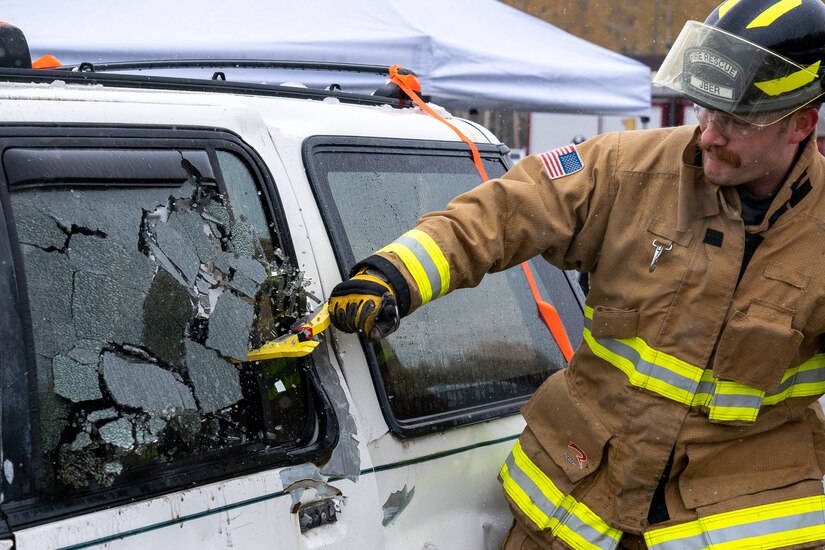 Nicholas Parks, a 673d Civil Engineer Squadron firefighter, uses a multitool to break the window of a damaged car for a vehicle-extrication demonstration during Fire Prevention Day.