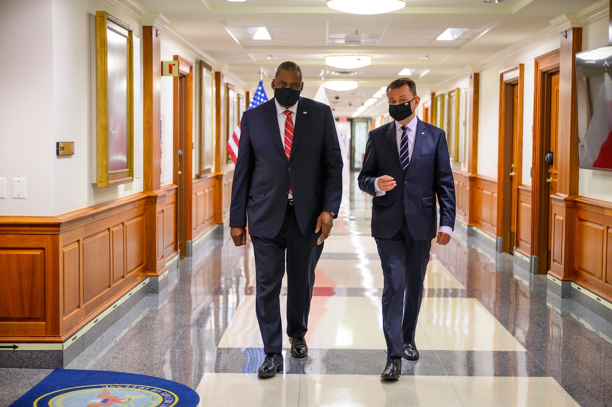 Two men walk down a hallway toward the camera. There is an American flag in the background.